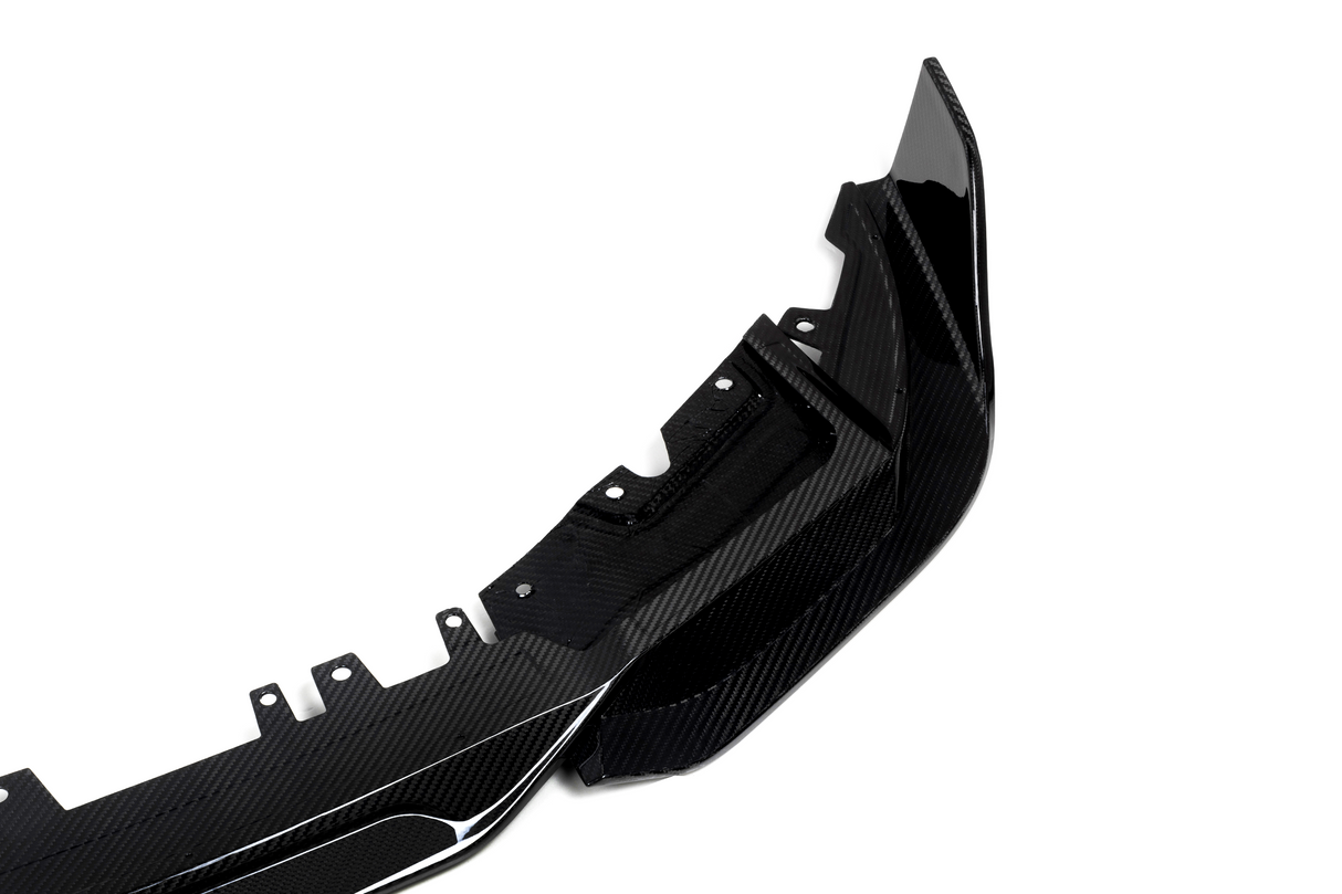 A glossy carbon fiber car accessory, likely a bumper, lies on a plain white background. The item features aerodynamic curves and multiple mounting points for attachment.