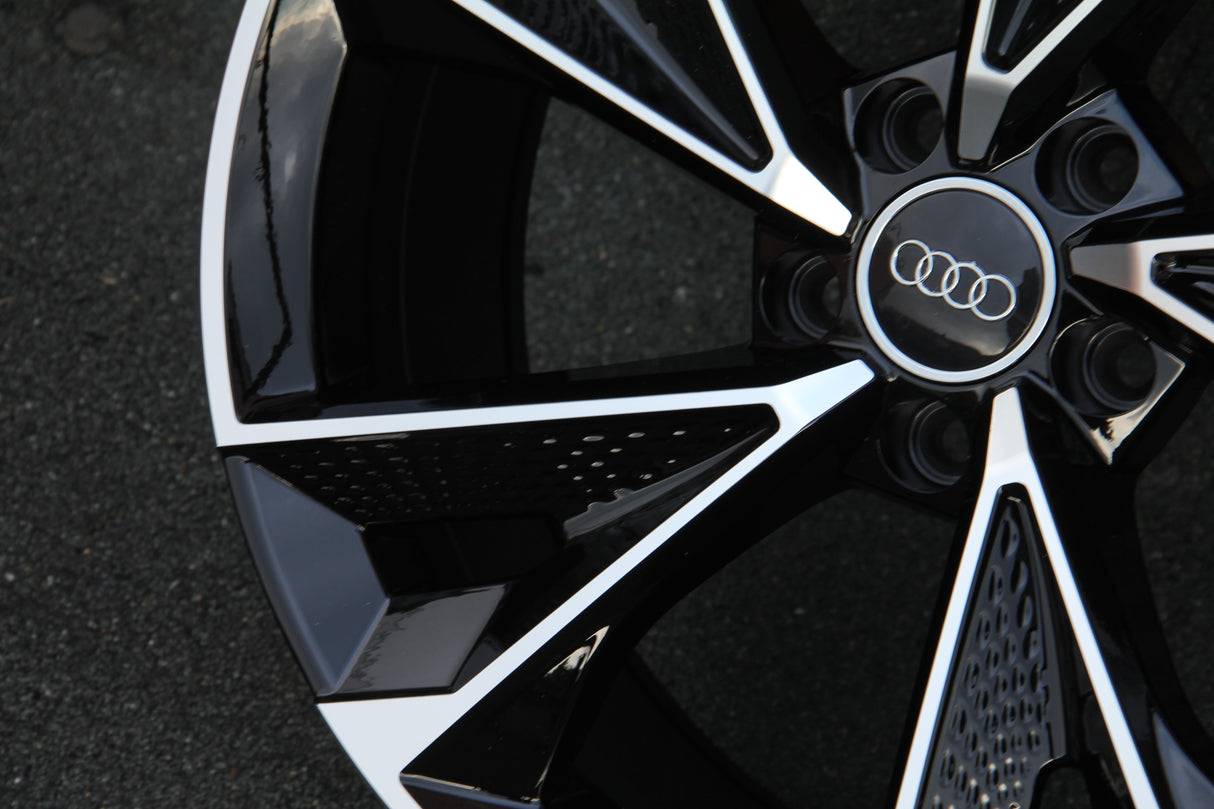 A shiny black and silver car wheel rim with the Audi logo at the center, positioned on a dark, textured ground surface.