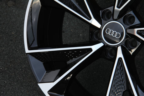 A black and silver Audi wheel rim with a logo centered; sunlit reflections create contrast, resting on textured asphalt pavement.