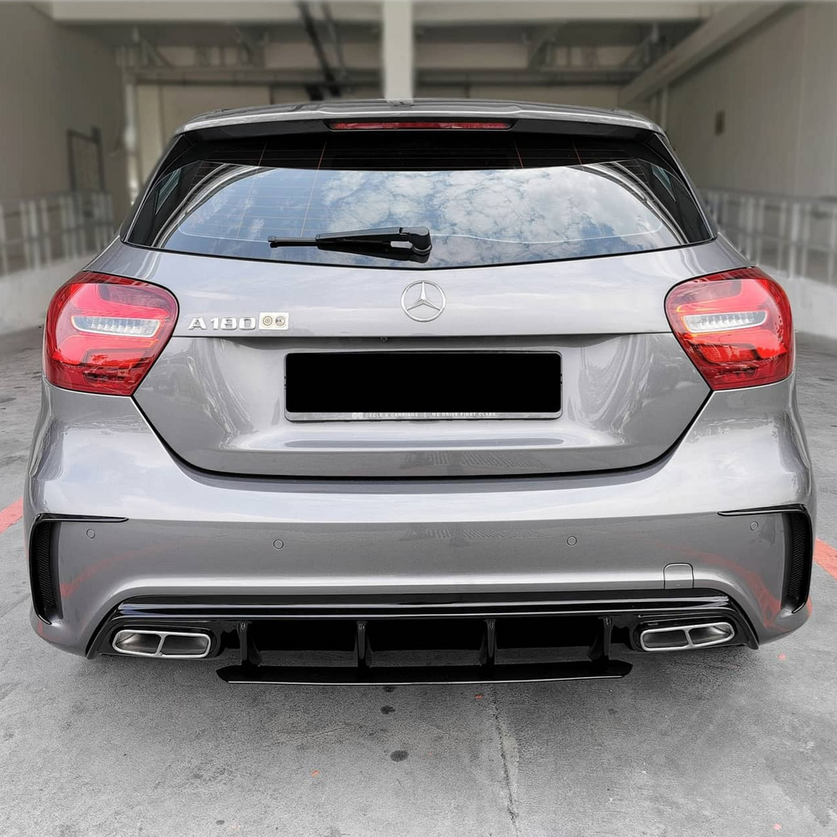 A gray Mercedes-Benz A180 car is parked, with visible rear lights and dual exhausts, in a concrete parking area. The badge "A 180" is on the left.