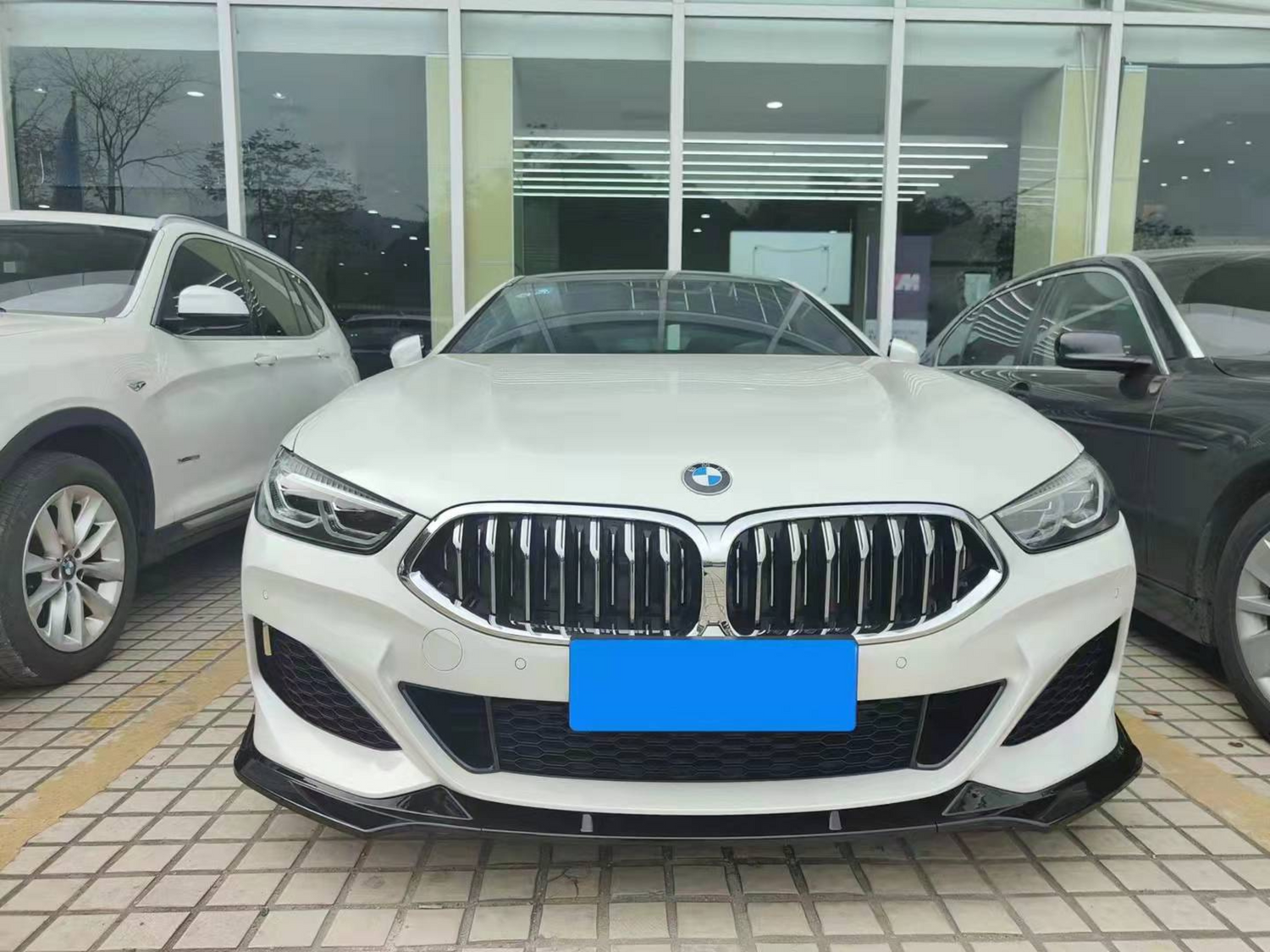 White BMW parked between two vehicles in front of a glass-fronted building. The car's front grille and headlights are prominently displayed on a tiled surface.