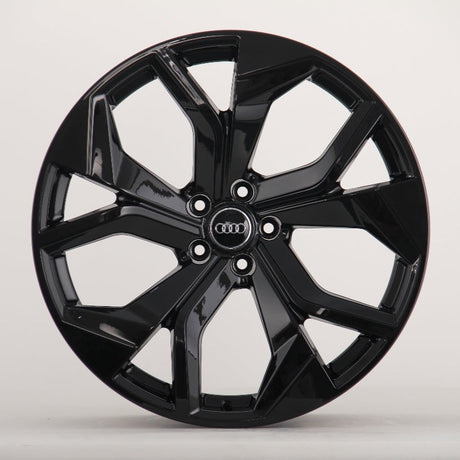 A glossy black alloy wheel with a multi-spoke design. The Audi logo is centered on the hub. It's set against a plain, light grey background.