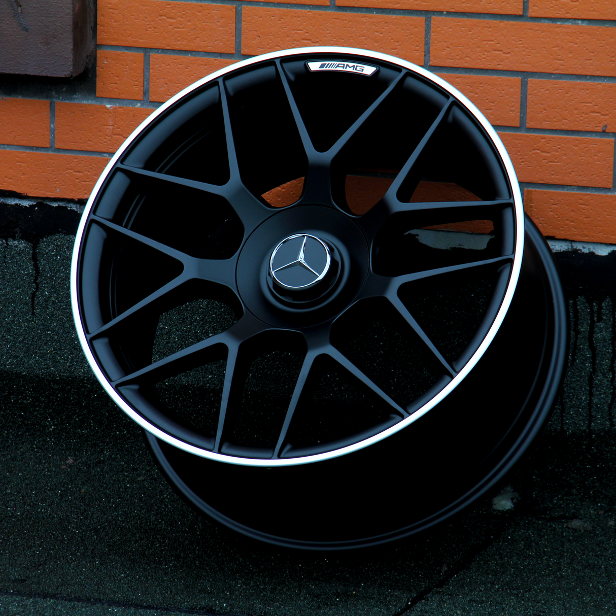A black AMG car wheel rests against an orange brick wall. The wheel features a Mercedes logo at its center.