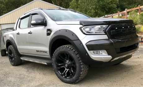 A silver pickup truck with "WILDTRAK" on the side is parked on a gravel driveway beside a building, surrounded by greenery. The front grille displays the "FORD" logo prominently.