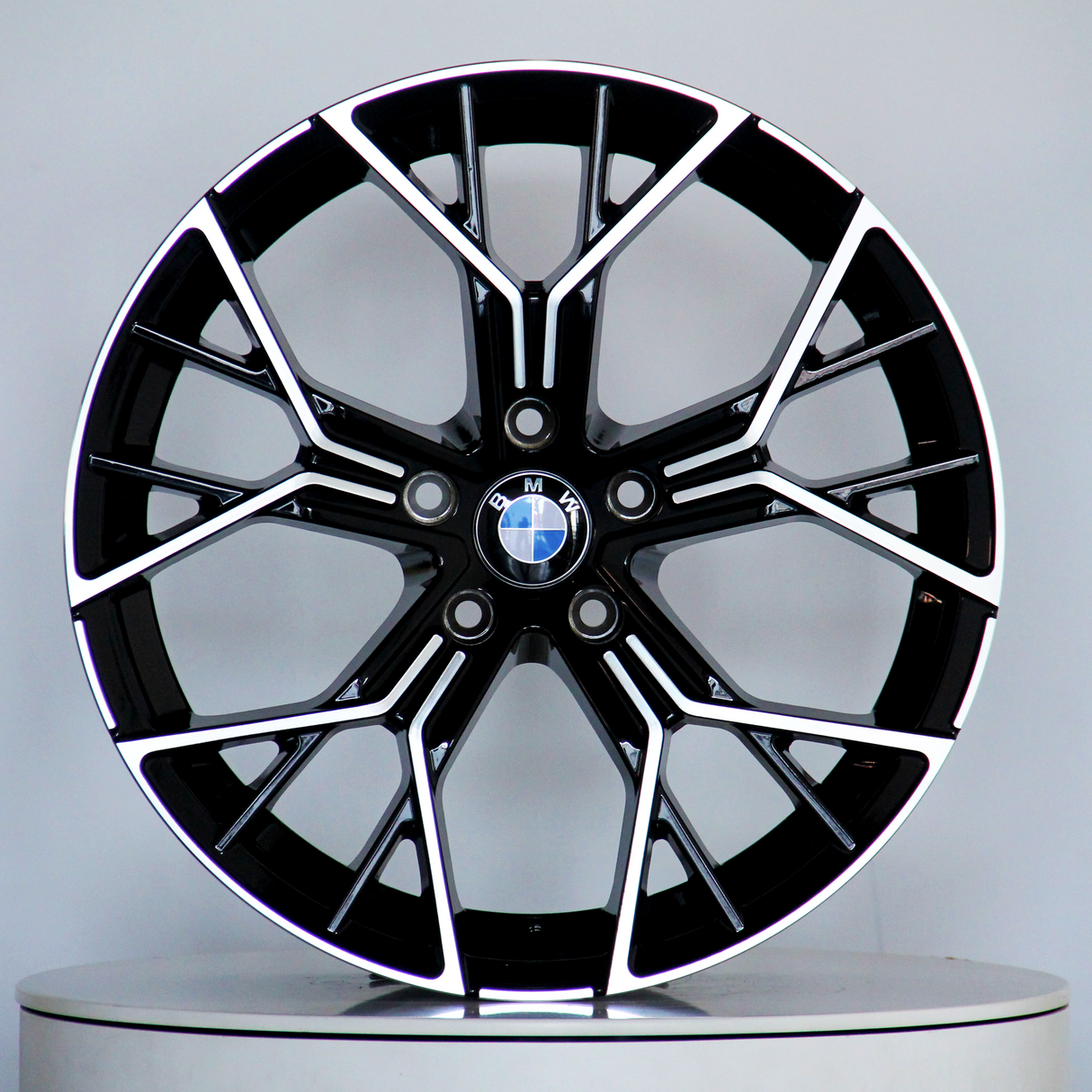 A black and silver BMW alloy wheel, featuring a multi-spoke design, is displayed upright on a light gray circular platform. The BMW logo is centered.