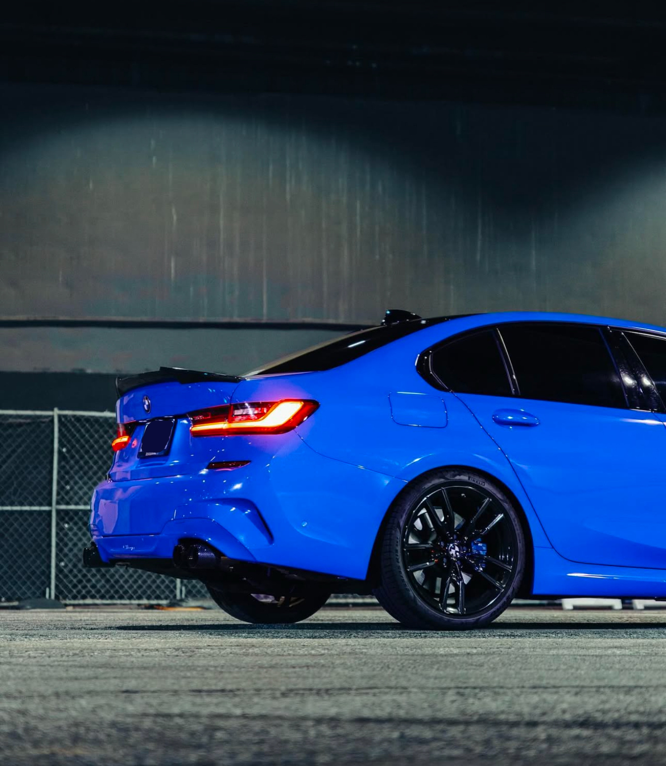 A blue sedan with illuminated taillights is parked on a concrete surface, surrounded by a chain-link fence and under an industrial-style structure.