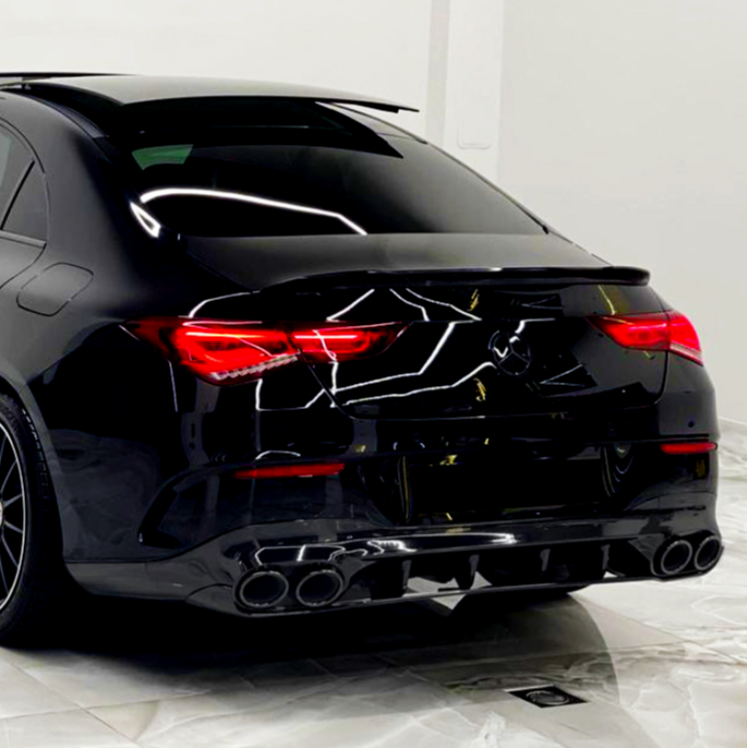 A sleek, black car with illuminated red taillights is parked on a marble floor, reflecting geometric ceiling lights in a pristine, white-walled room.
