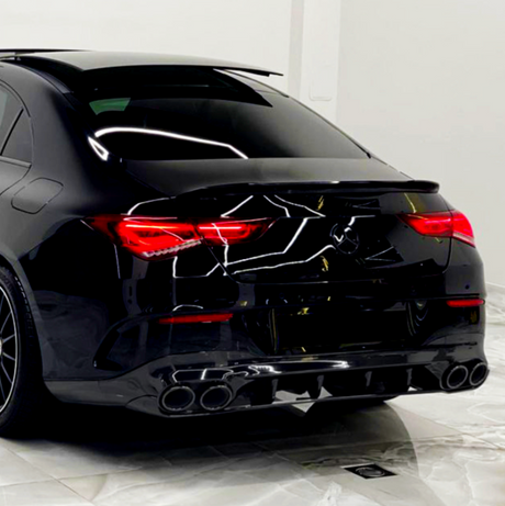 A sleek, black car with illuminated red taillights is parked on a marble floor, reflecting geometric ceiling lights in a pristine, white-walled room.