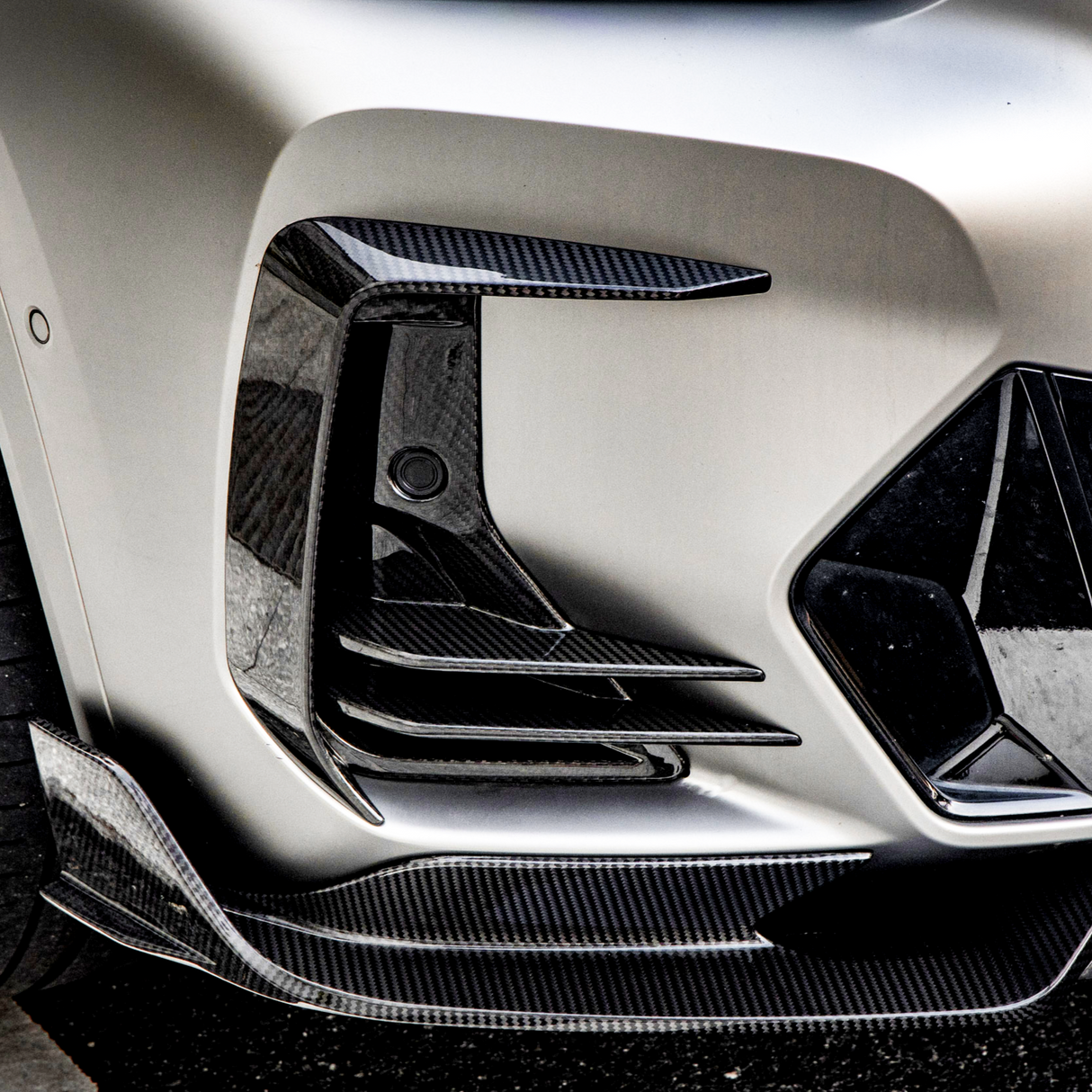 A carbon fiber car bumper features intricate aerodynamic vents and sleek curves, set against a smooth, metallic body, resting on textured asphalt.