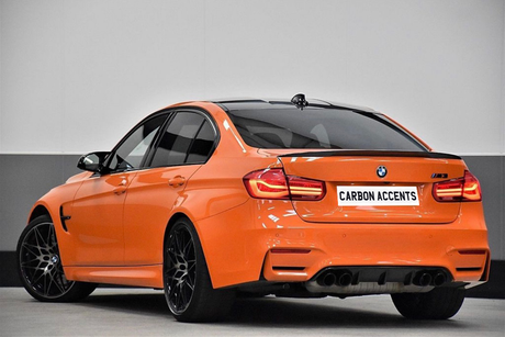An orange BMW M3 is parked indoors, showcasing a sleek design and black wheels. The license plate reads "CARBON ACCENTS," emphasizing its sporty appeal in a neutral setting.