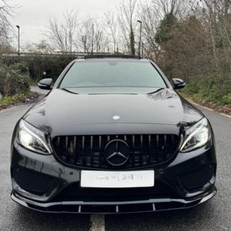 A black Mercedes car is parked on a road lined with leafless trees and bushes. It has illuminated headlights and a visible Mercedes emblem at the front center.