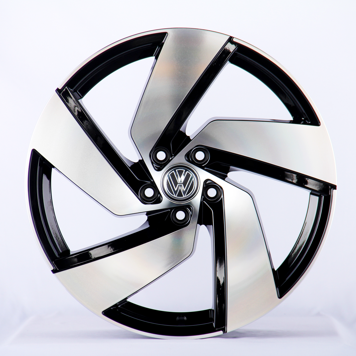 A sleek, silver and black alloy wheel with an intricate design sits upright in a well-lit studio. The central emblem features the Volkswagen logo against a plain white background.