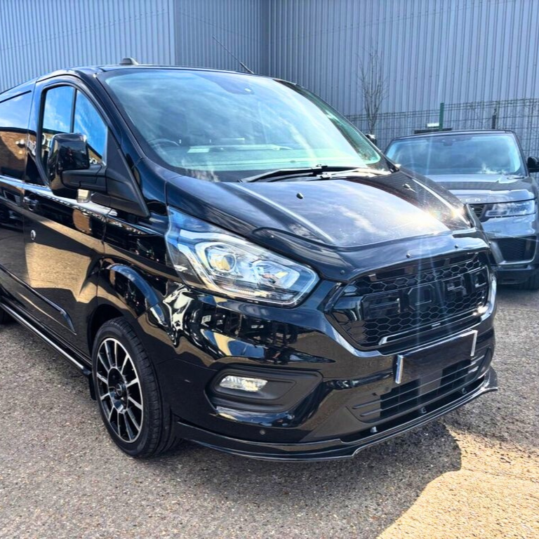 A black van is parked on a paved surface beside another vehicle, in front of a large industrial building under a clear sky.