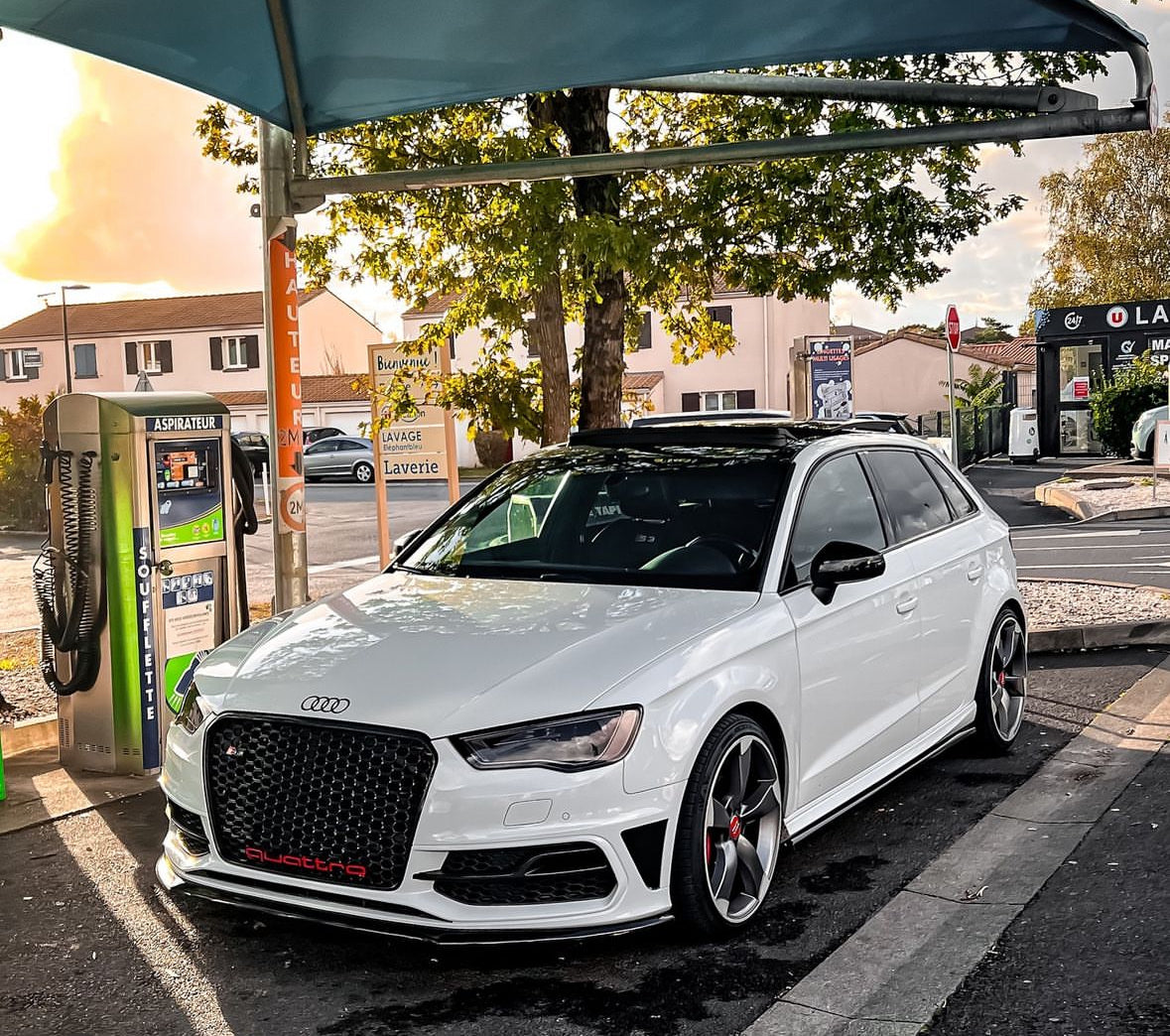 A white Audi car is parked beside a vacuum machine under a canopy in a car wash area. Nearby, signs read “Aspirateur,” “Soufflette,” “Bienvenue,” “Lavage,” “Laverie.” Houses and trees are visible.