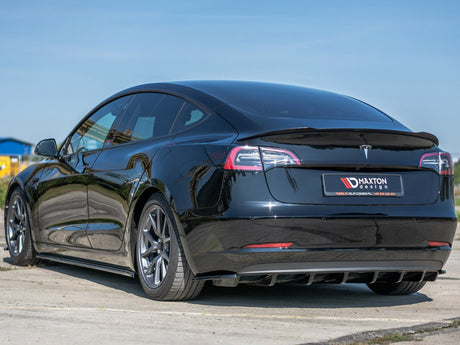 A black car parked on a pavement, showcasing its sleek design with tinted windows and alloy wheels. The background includes a clear blue sky and distant buildings. License plate reads: Maxton Design.