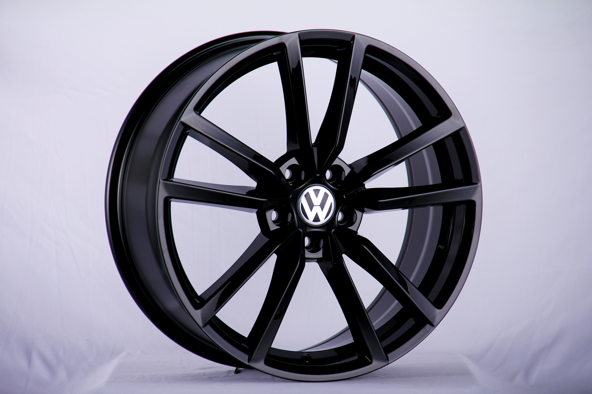 A black alloy wheel with a Volkswagen logo in the center, standing upright on a plain, light-colored background.