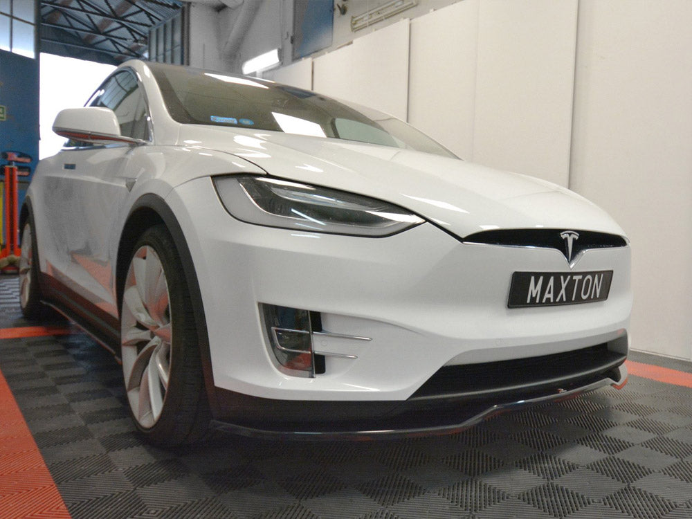 A white car with a Tesla logo is parked indoors on a checkered floor, displaying the license plate "MAXTON." The room features a high ceiling and bright lighting.