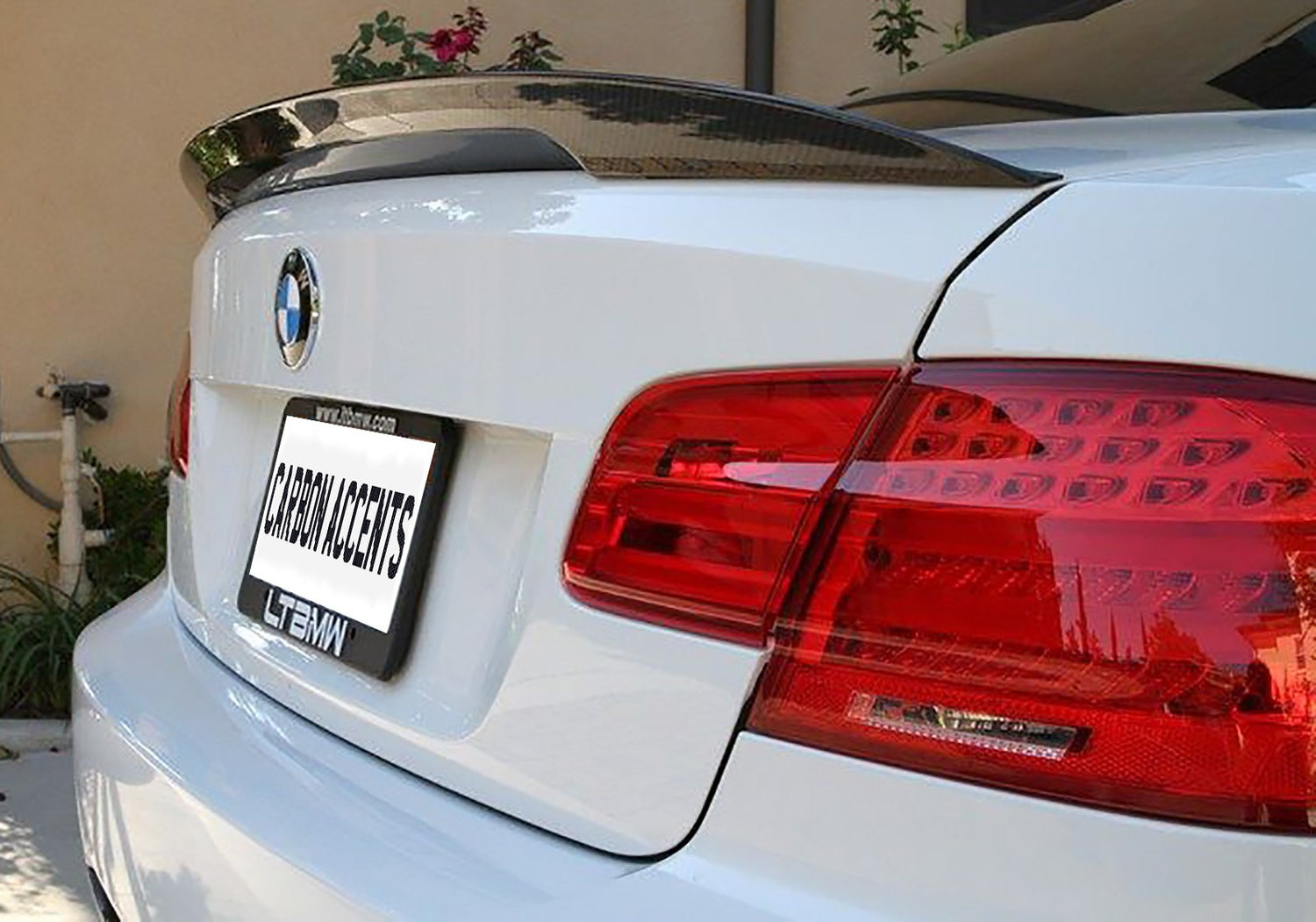 A white car features a carbon fiber spoiler and red tail lights. The rear license plate shows the text "CARBON ACCENTS." It's parked in a driveway with plants nearby.