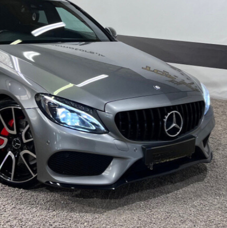 A silver luxury car with illuminated headlights is parked indoors on a textured carpet. The background features a white wall with a black stripe. No text is visible.