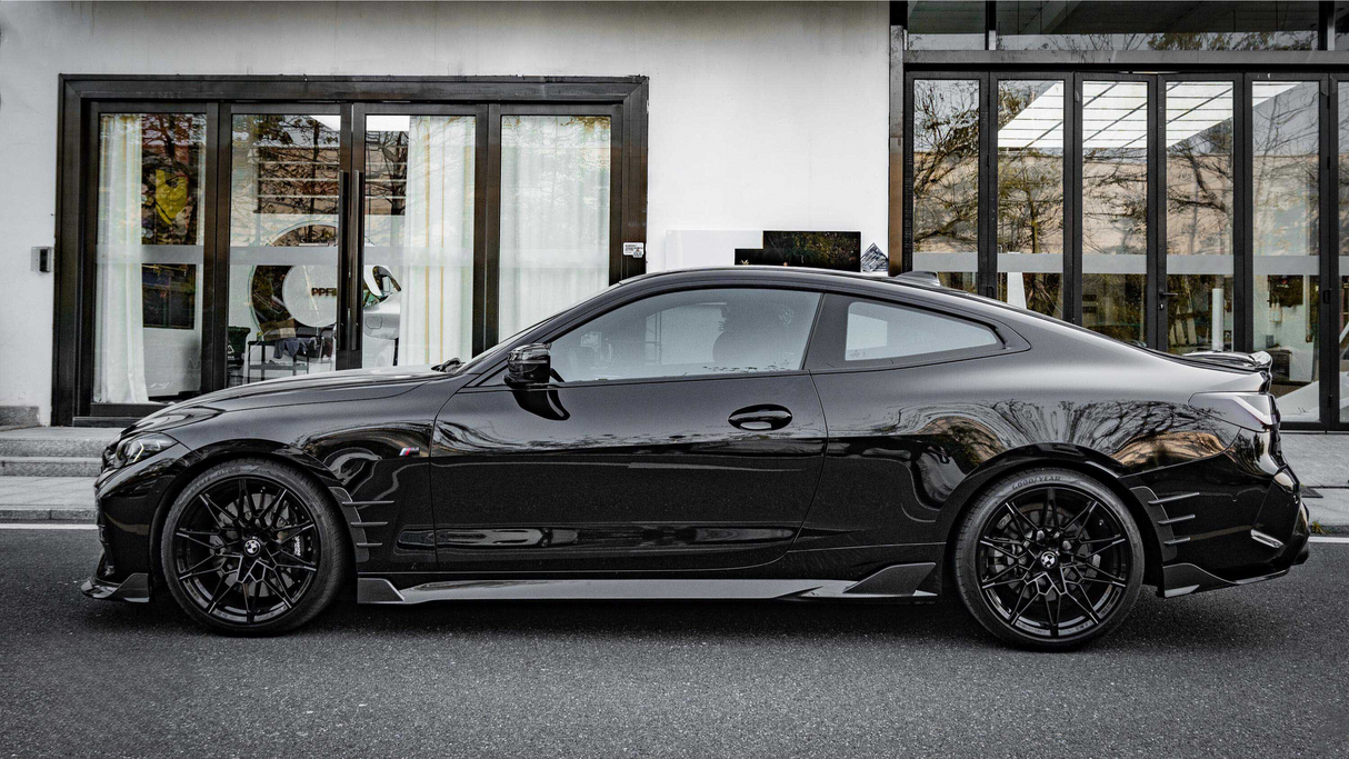 A sleek black sports coupe is parked in front of a modern building with large glass doors and light curtains, reflecting the surrounding trees and architecture.
