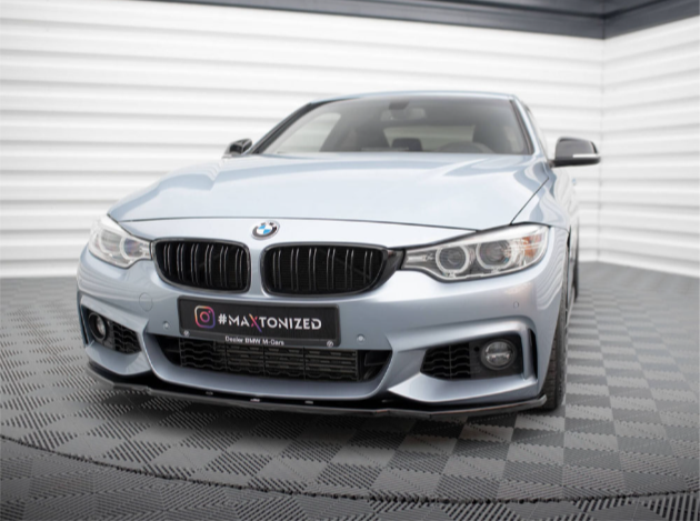 A silver BMW car is parked indoors, facing forward on a checkered floor. The license plate reads "#MAXTONIZED, Dealer BMW M Góra." The room has striped white and gray walls.