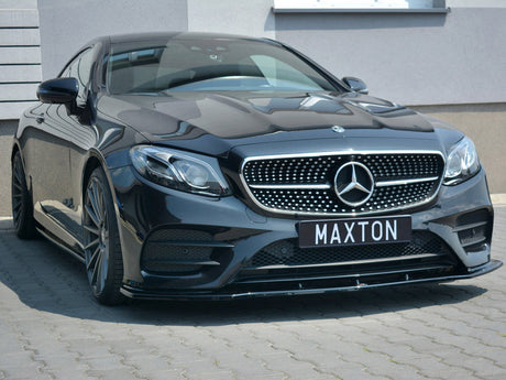 A black Mercedes-Benz car is parked on a paved surface, featuring a prominent grille with a star emblem. The license plate reads "MAXTON," and it's beside a modern building.
