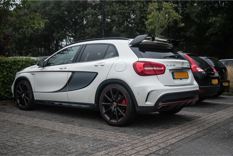 A white car with a sporty design, labeled "GLA 45" and black-and-red accents, is parked on a paved road. Other vehicles and leafy bushes are in the background.
