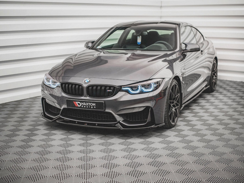 A sleek, gray BMW sports car with illuminated headlights sits on a checkered floor in a bright, white-paneled room. A license plate reads: "MAXTON design."