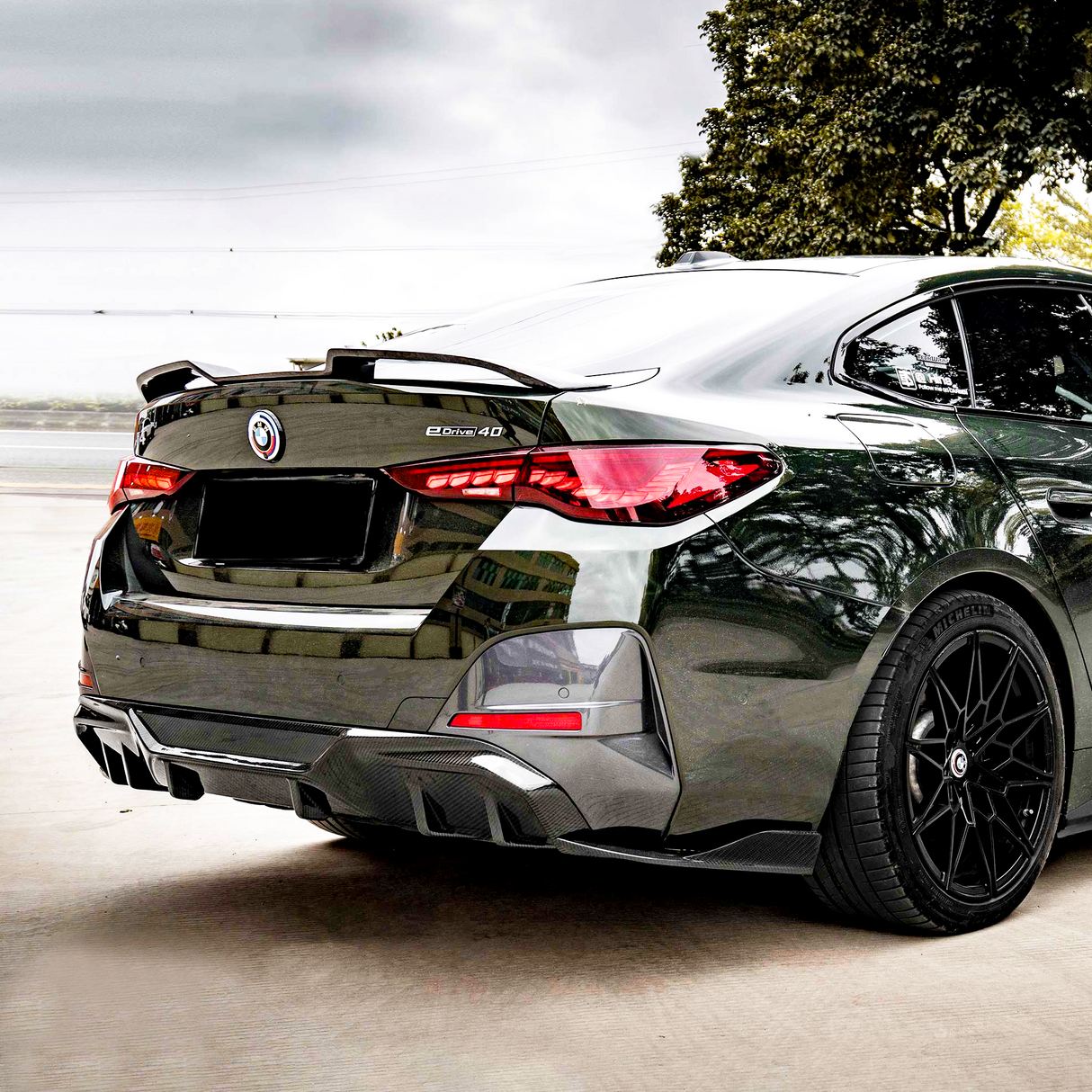 A sleek black car with a "b3x5 4D" badge is parked on a concrete surface, surrounded by trees and a cloudy sky.