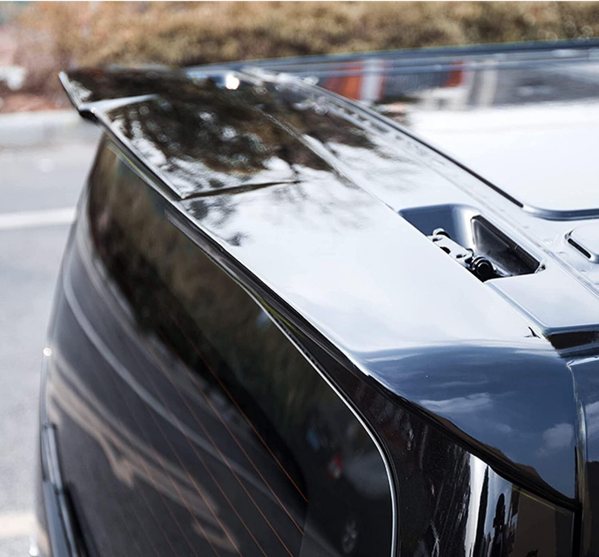 A sleek, glossy car spoiler is installed on the rear of a black vehicle, reflecting trees and sky, situated in an outdoor parking area.