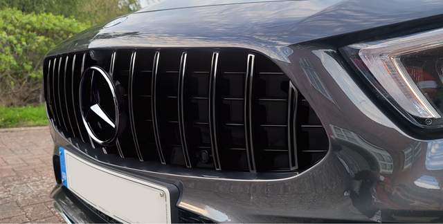 Car grille with vertical slats featuring a circular emblem in the center, situated on a parked dark-colored vehicle; surrounded by a paved area and greenery in the background.