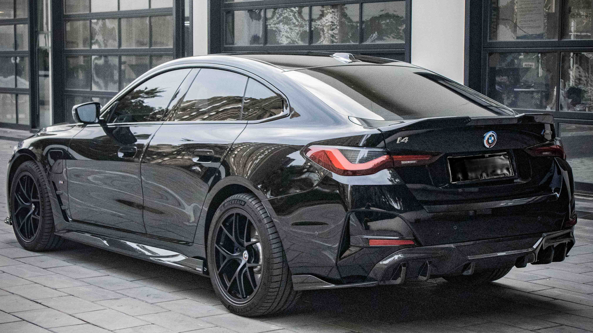 A black BMW i4 parked on a cobblestone driveway outside a modern building with large windows. The car's design features sleek lines and tinted windows, with badges displaying "BMW," "i4," and "M50".