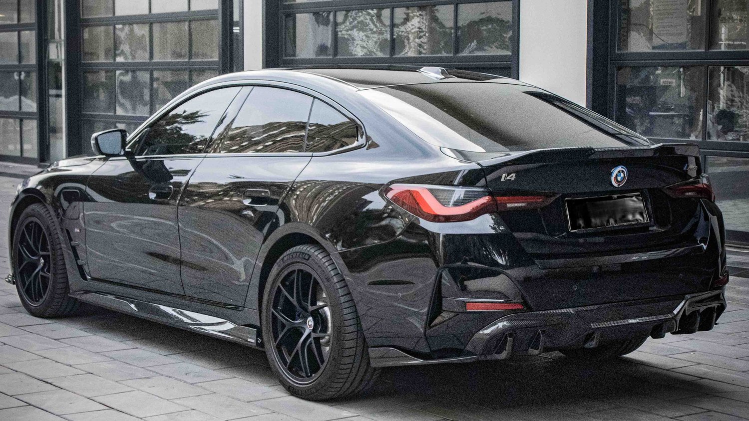 A black BMW i4 parked on a cobblestone driveway outside a modern building with large windows. The car's design features sleek lines and tinted windows, with badges displaying "BMW," "i4," and "M50".