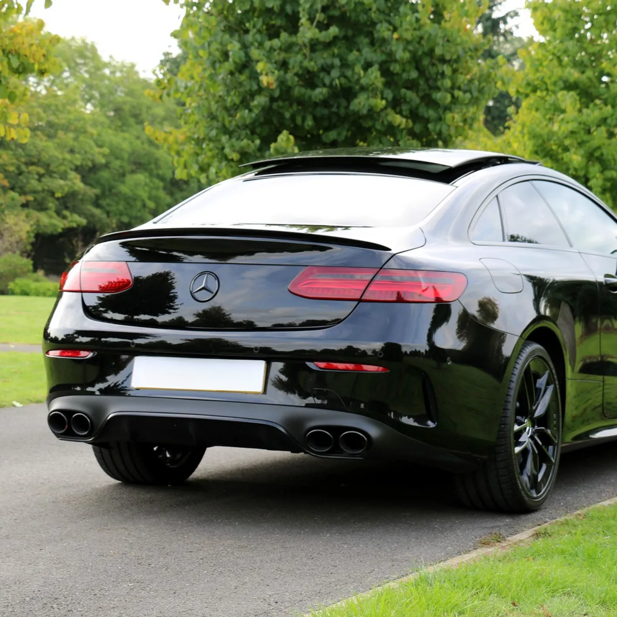A black car with a Mercedes logo parked on a road, surrounded by greenery and trees. The car features tinted windows and dual exhaust pipes, positioned in a park-like setting.
