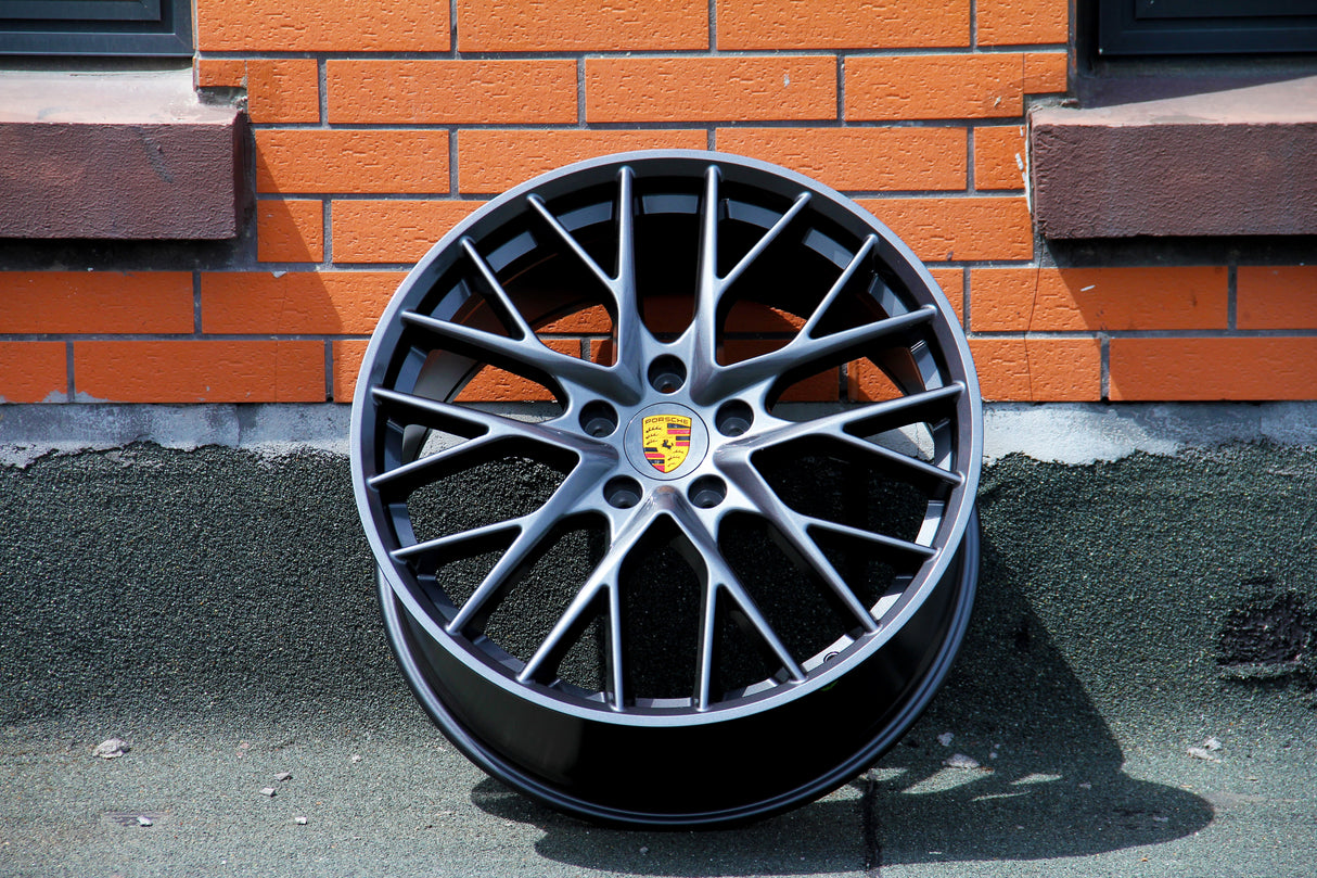 A car wheel rim with multiple spokes, displaying the Porsche logo, leans against an orange brick wall on a rough, paved surface.