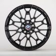 The image shows a black alloy wheel with a complex geometric design, featuring a blue, white, and red BMW logo at the center. The wheel is set against a plain white background.
