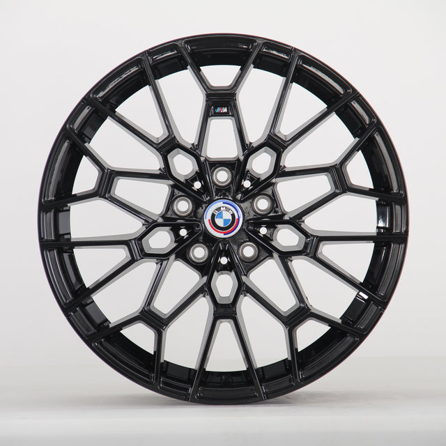 The image shows a black alloy wheel with a complex geometric design, featuring a blue, white, and red BMW logo at the center. The wheel is set against a plain white background.