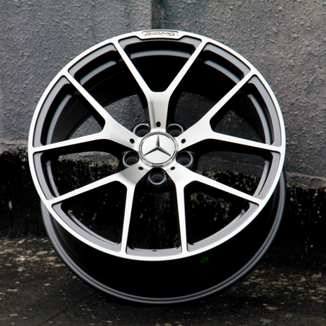 A metallic car wheel with a Mercedes-Benz logo and "AMG" text rests upright against a gray textured wall, situated on a rough pavement surface.