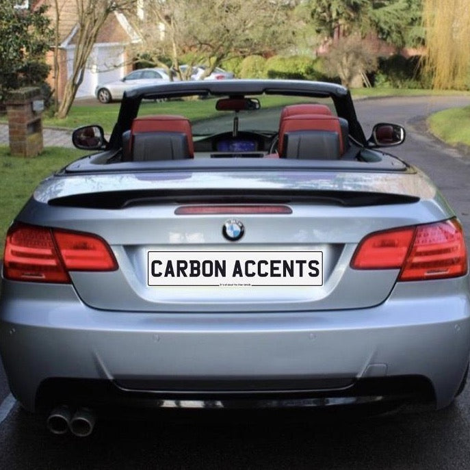 Convertible silver car with red interior parked on a residential street. License plate reads "CARBON ACCENTS." Brick mailbox and house in the background surrounded by trees and grass.
