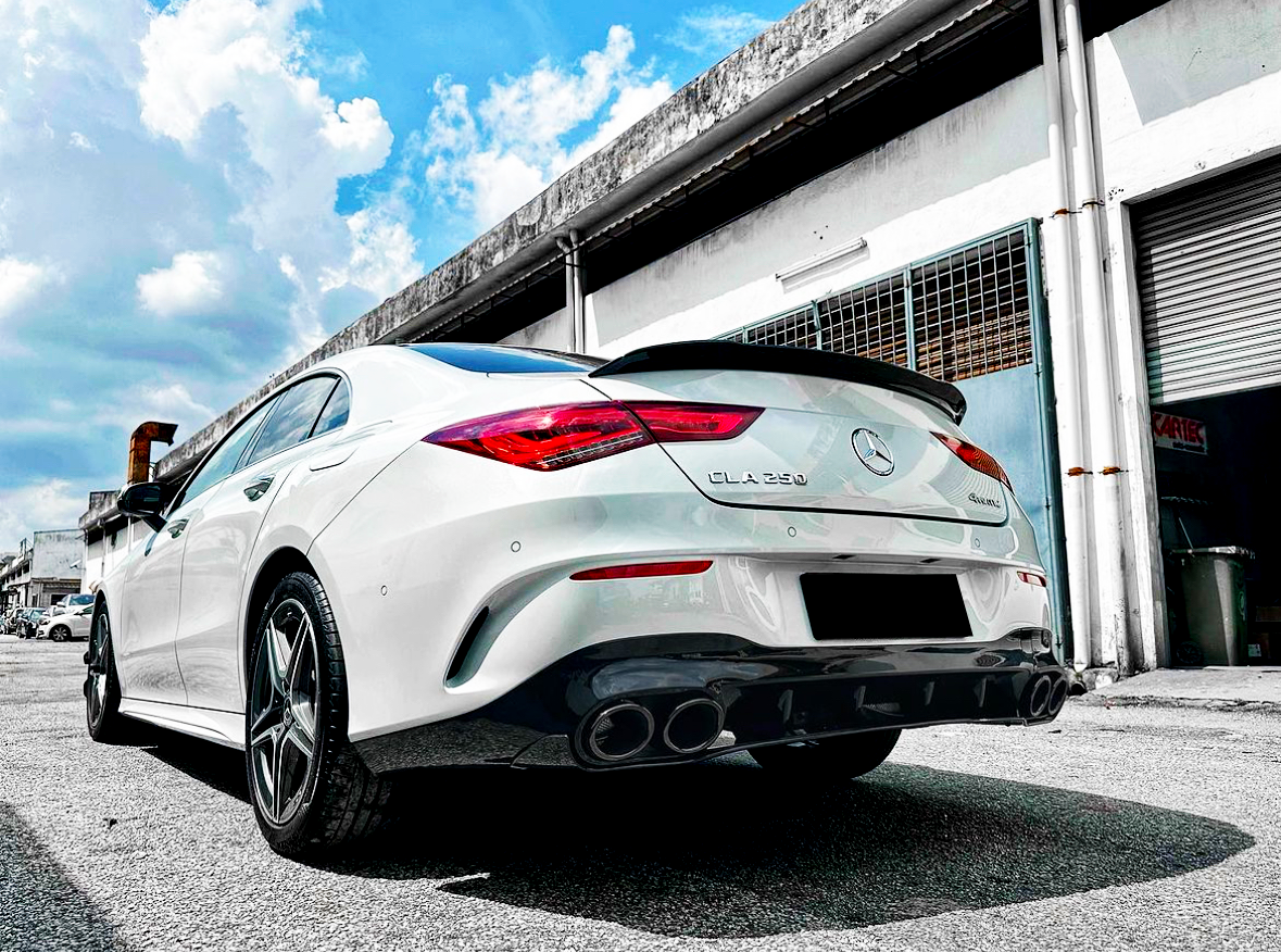 A white Mercedes-Benz CLA 250 faces away on a sunny day in an industrial area, parked near a building with a partially open garage door.