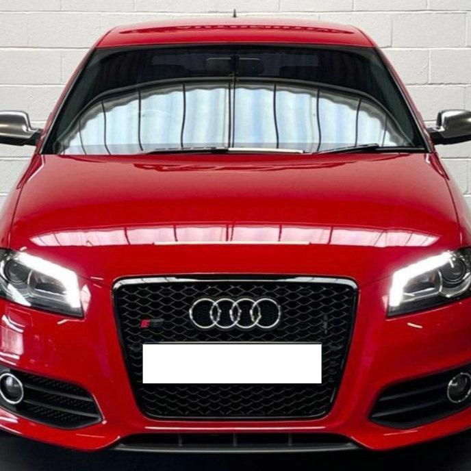 A red Audi car, stationary, with headlights on, is parked indoors against a white brick wall. The car displays a black grille with the Audi logo and a blank license plate.