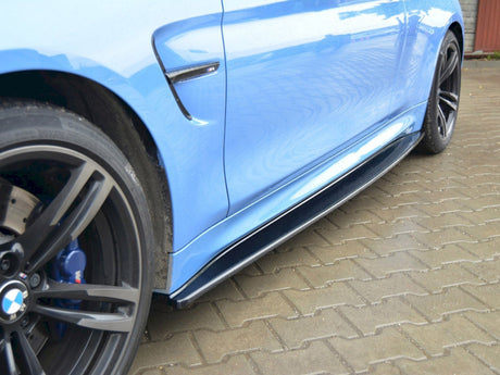 A close-up of a blue sports car's side, focusing on the black side skirt and alloy wheels, parked on interlocking brick pavement. The BMW logo is visible on the wheel.