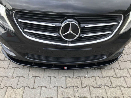 A black car front features a prominent emblem in the center of a chrome grille, parked on a paved surface with interlocking bricks. The license plate area is empty.