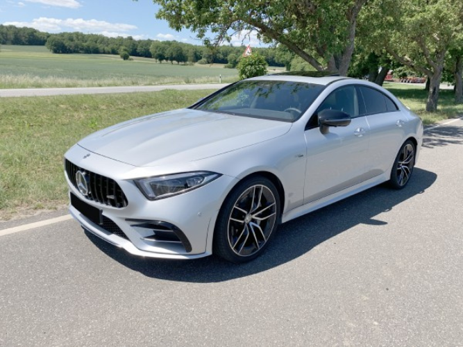 A sleek silver car is parked on a sunny road beside grassy fields and trees. It features black alloy wheels and a modern aerodynamic design.