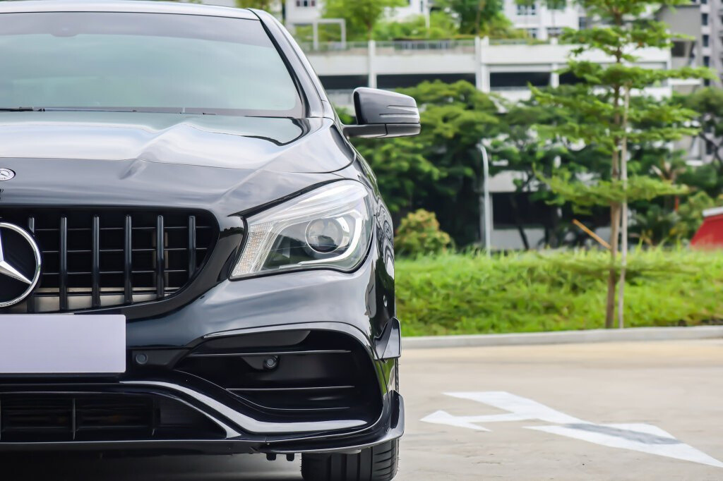A black car's front view with a shiny grille and headlight is parked near green foliage and buildings in the background.