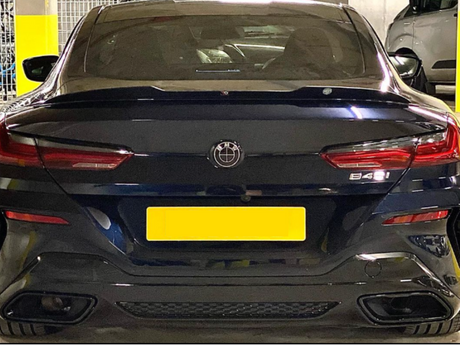 A black car parked in a garage displays a yellow license plate and badge. The model marked is "B48I". Surrounding are other vehicles and a concrete wall.