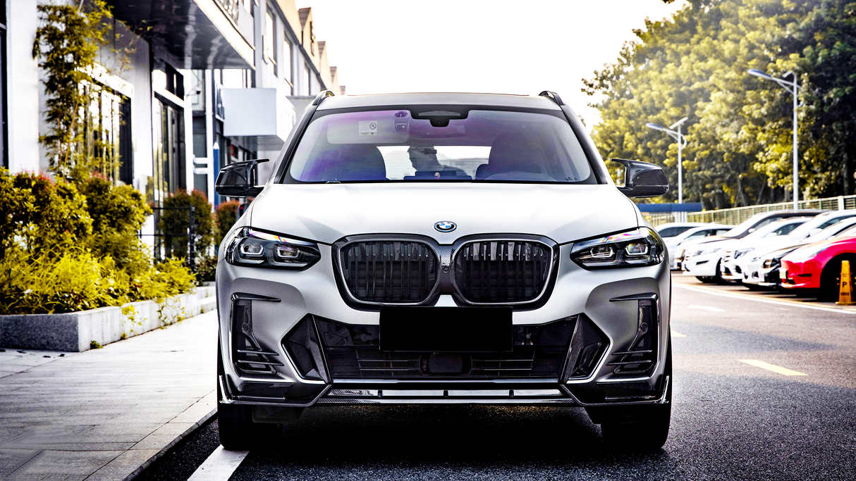 A white car is parked on a street, facing forward. It is surrounded by parked cars and trees line the side of a nearby building.