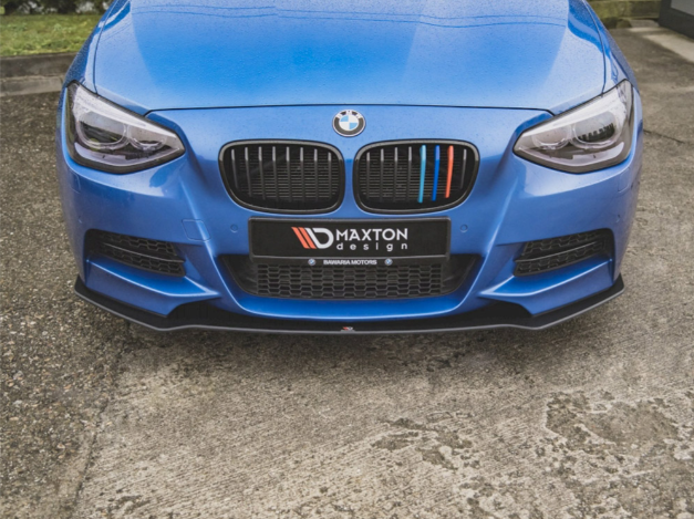 A blue BMW car front displays a black grille with colorful stripes; "MAXTON design" text is on the license plate cover. The car is parked on a concrete surface.