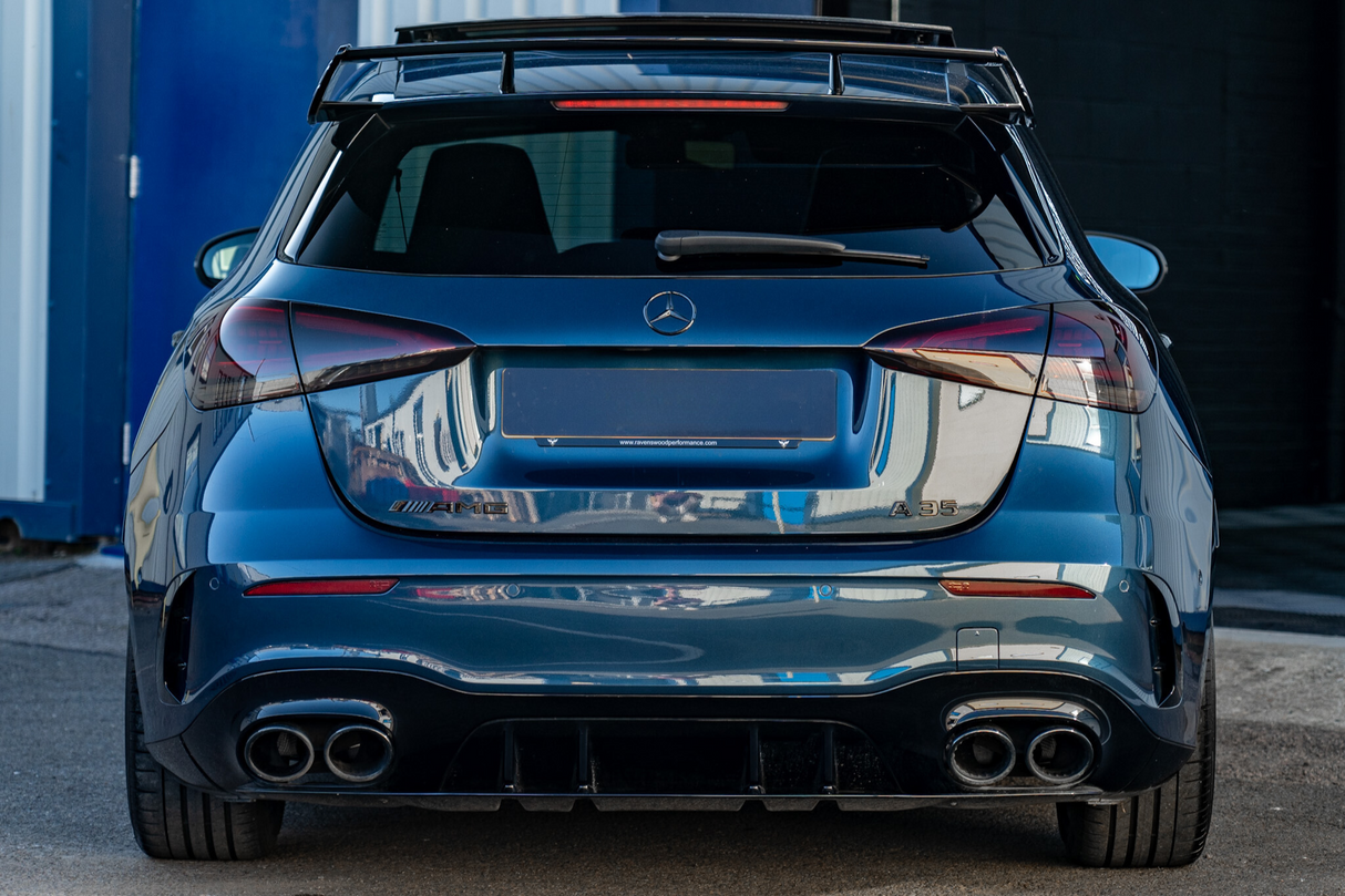 Blue Mercedes AMG A35 parked, displaying dual exhausts and a spoiler. It is situated in an industrial area, surrounded by buildings. Text on the license plate reads: "www.reveriecooperformance.com".