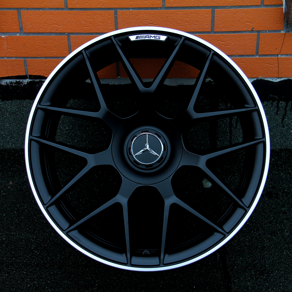 A black Mercedes-Benz AMG wheel rim rests against an orange brick wall. The central logo features a three-pointed star, with "AMG" written on a small badge near the top.